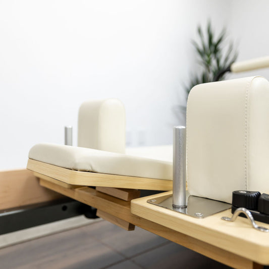 Reformer Pilates machine with cream upholstery and wooden frame.